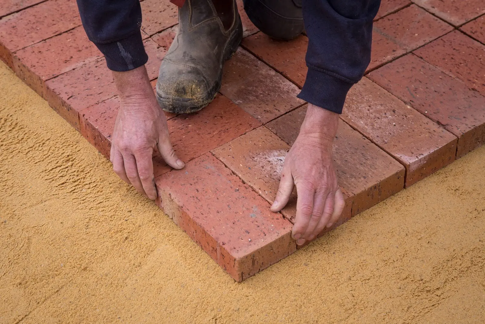 Hands setting red brick pavers into a leveled sand base